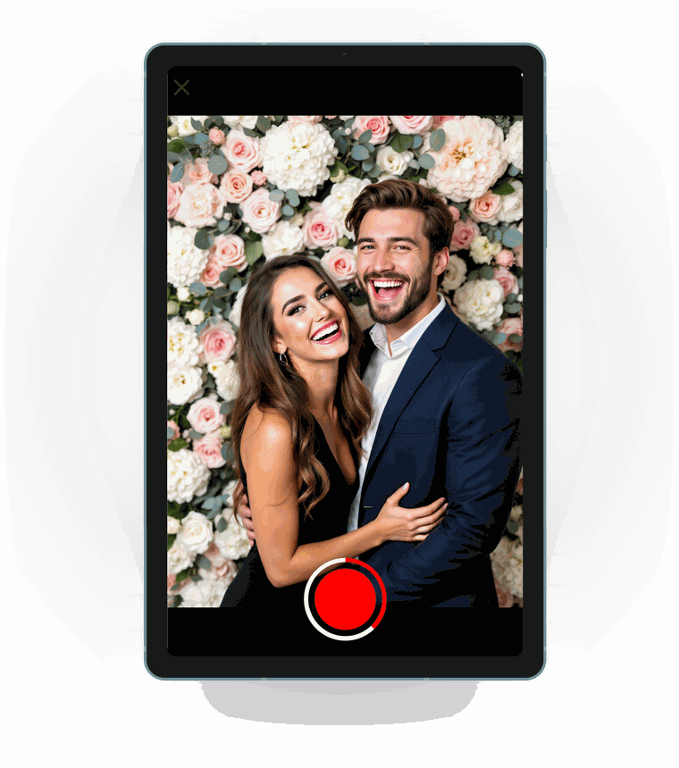 Android tablet running LumaBooth as a photo booth, a young couple in evening wear laughing together in the live camera view in front of a white and blush pink flower wall, with the capture button below